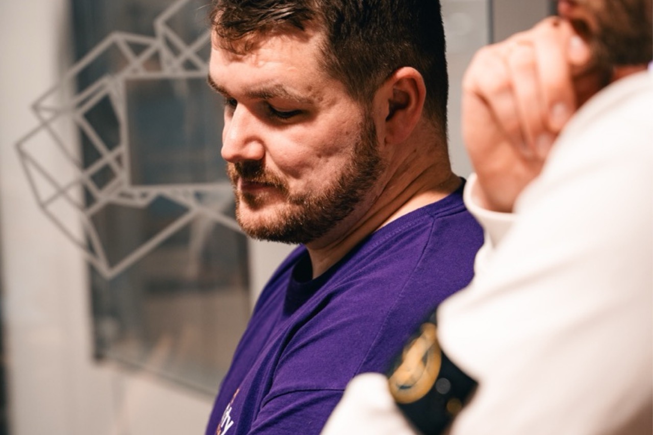 Mann im violetten Shirt scharf im Bild; eine zweite Person mit heller Kleidung liegt unscharf im Vordergrund