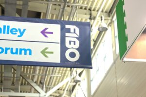 Deckenleuchten und Tageslicht über einem FIBO-Wegweiser in einer Messehalle