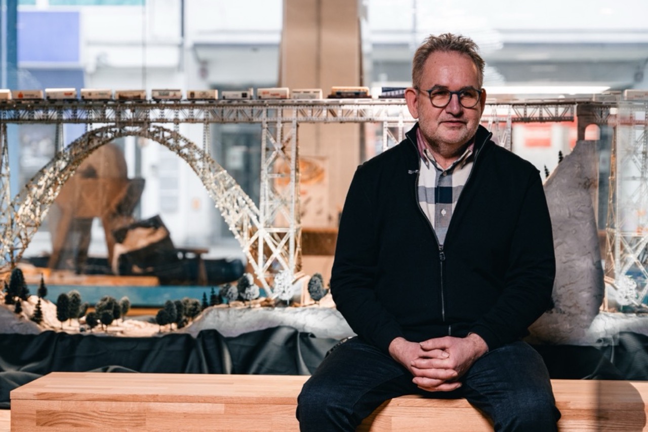 Sitzender Mann mit Brille vor einer Modellbrücke und mehreren Modellfahrzeugen