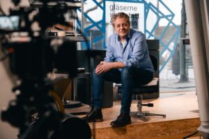 Mann im blauen Hemd sitzt für eine Interviewaufnahme vor einer Kamera neben einer Glasfront
